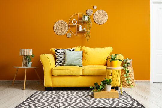 Comfortable yellow sofa and stylish tables with houseplants near orange wall in living room