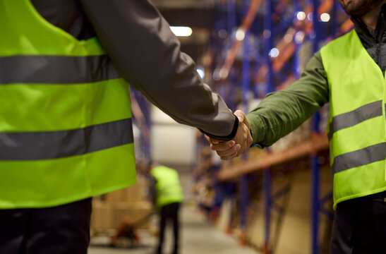 Handshake workers warehouse logistics delivery teamwork agreement. Staff in safety vests confirm inventory and shipping details on site. Symbol of partnership in warehouse operations.