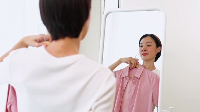 Cheerful Asian female holding hangers with casual shirts, preparing her outfit in a mirror. Concept of personal style development, dressing up and modern lifestyle.