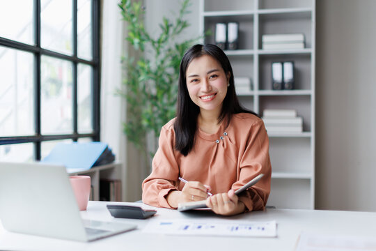 Young asian businesswoman smiling, working on financial data and planning notes at modern office desk, managing business tasks for career success and productivity