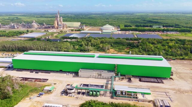 Large green industrial building marks Ecoacero steel rod factory in Dominican Republic, solar panel array plus distant cement plant spread across surrounding cleared land. Aerial drone wide shot