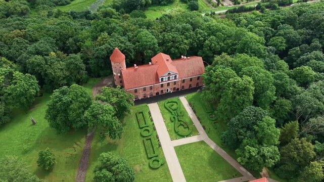 Stunning drone video captures Raudondvaris Castle and manicured baroque gardens, framed by lush trees. Clear sunlight highlights the red roof and greenery in this cinematic aerial footage.