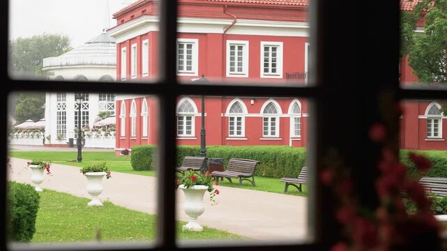 Soft natural daylight illuminates a peaceful courtyard at Raudondvaris Castle, Lithuania, framed by a window with vivid red architecture, green hedges, benches, and elegant white planters.