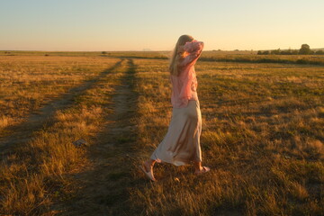Obraz premium Graceful blond woman in a silk skirt walking along a scenic dirt road in the steppe of East Kazakhstan at sunset. Elegant romantic fashion photography, back view.