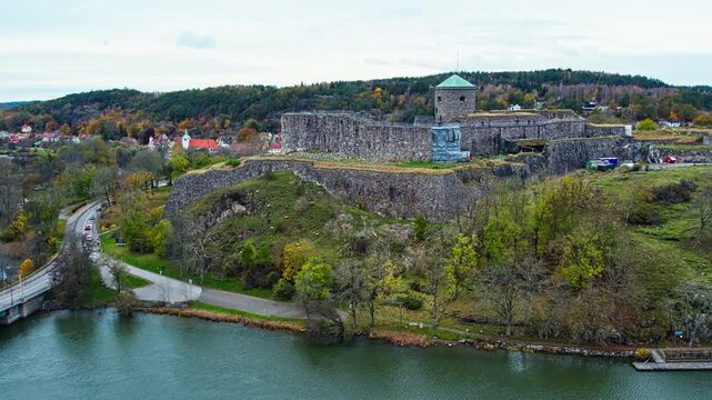 Scenic aerial drone view of the historic Bohus Fortress situated on a hill overlooking the river in Kung&auml;lv, Sweden. The stone ruins are surrounded by lush autumn trees under a cloudy sky.
