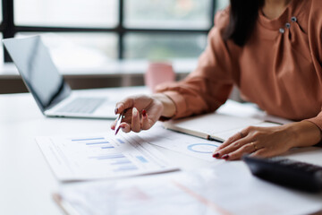 Plakat Professional woman analyzing financial data and business reports, using a pen to point at charts while working on a laptop at an office desk