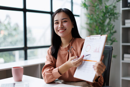 Young asian businesswoman smiling, presenting financial data on a clipboard in a modern office, showing progress and successful business analytics during a professional meeting