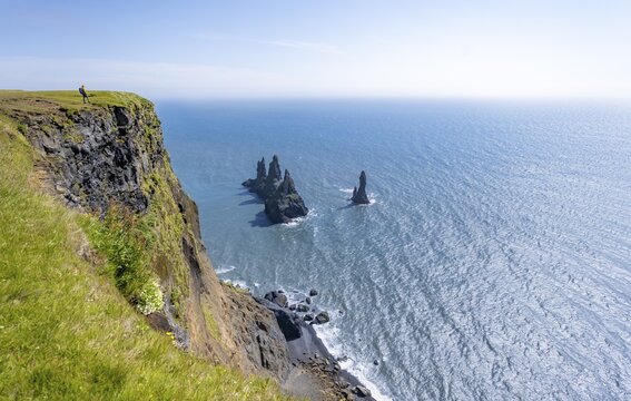 Young man standing on cliff, view from steep cliff to the rocks Reynisdrangar in the water, at Reynisfjara beach, Vik, South Iceland, Iceland