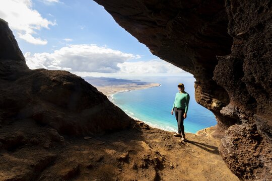 Tourist in the Cueva de las Cabras cave, young woman enjoying the view from the Risco de Famara cliff to Famara beach, Playa de Famara with La Calaeta, Lanzarote, Canary Islands, Spain