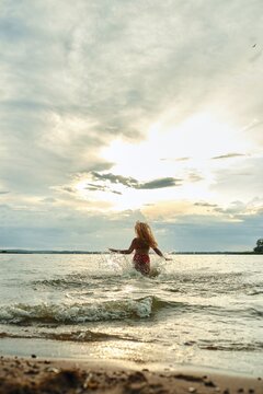 Funny woman plays joyfully in the water at a lakeside beach. The sun sets behind her, casting beautiful colors in the sky. The atmosphere is cheerful and serene as she splashes water around