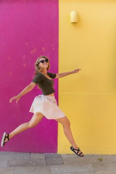 Young woman in dress jumps happily in front of colorful house, yellow and pink house facade, Burano Island, Venice, Veneto, Italy