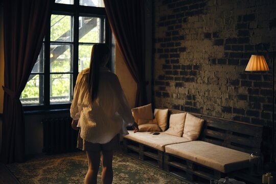 A woman stands in a softly lit room with brick walls and wooden accents. She wears a loose shirt and looks towards a window, where daylight illuminates the space, adding warmth to the atmosphere
