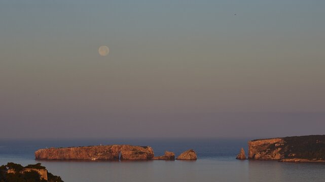 The moon is high above rocks and calm water, the sky is pink, Tsichli Baba island, Sfaktiria island, Navarino Bay, Pylos, Peloponnese, Greece