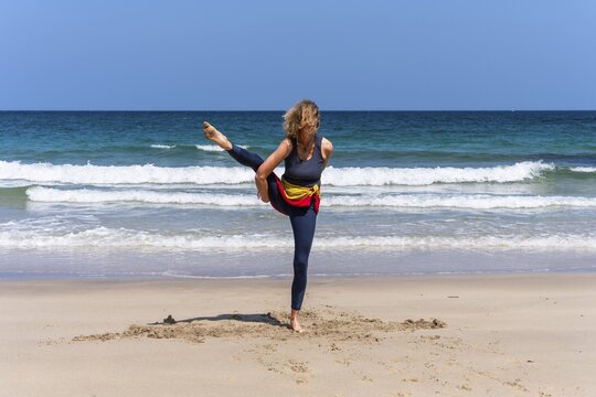 A woman doing yoga on a sandy beach by the sea. Svarga Dvijasana or bird of paradise. Treen Cove, Cornwall, England, Great Britain