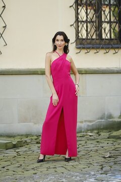 A woman poses confidently in a bright pink dress with a stylish slit, standing on a cobblestone path near an old brick building with decorative windows