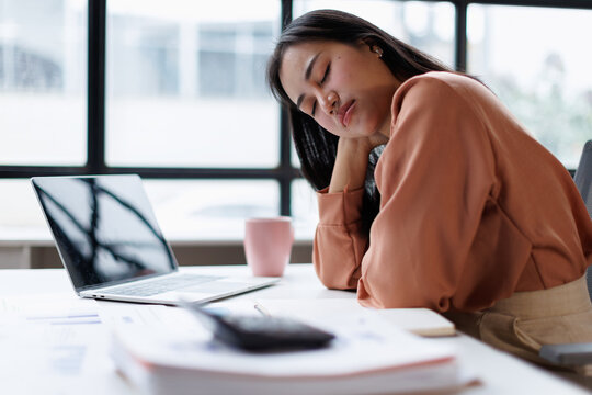Exhausted asian businesswoman sleeping at office desk, feeling fatigued and stressed from overwork, experiencing burnout, and needing a break from intense workload