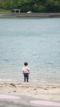 Small child stands at water's edge, barefoot. Waves lap gently at sand, soft breeze rustles hair. Child gazes out, curious, quiet, absorbed. Pink footprints mark path, leading to shoreline