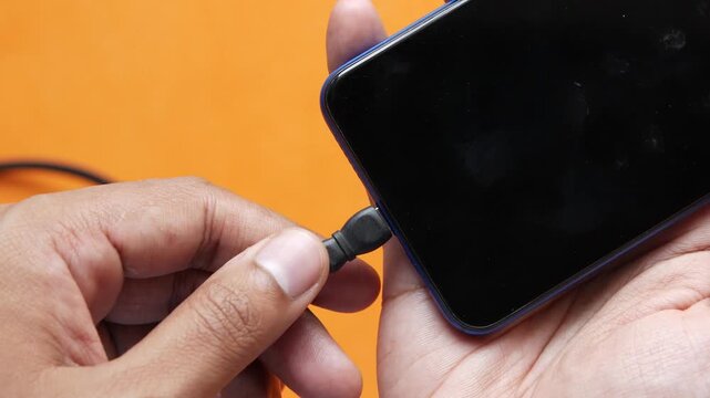 Person plugging micro usb charging cable into smartphone against orange background