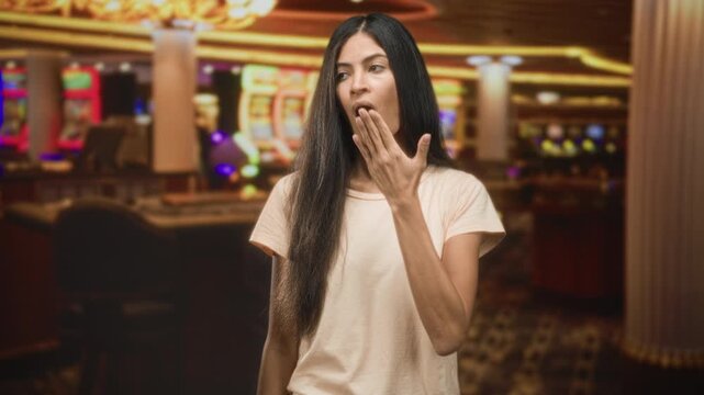 Woman covering mouth with hand yawns inside a casino building with slot machines and patterned carpet visible; fatigue waiting.