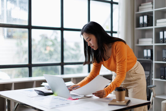 Young asian business woman analyzing financial data and working on startup marketing strategy, looking at documents and laptop screen in a modern office