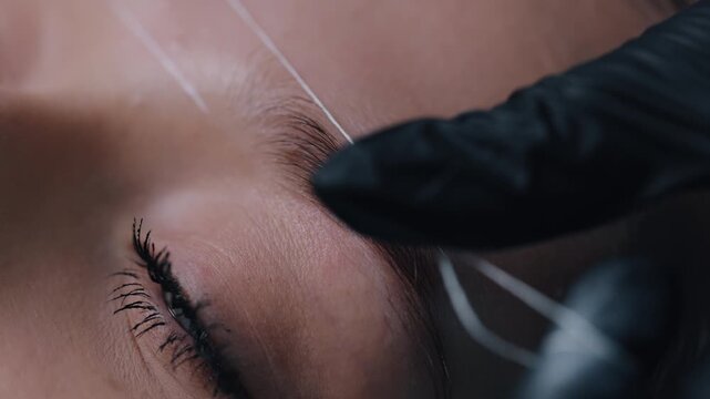 beauty salon, facial aesthetics, brow artist. Close-up beautician wearing black nitrile gloves performing eyebrow threading on young woman in professional beauty salon, emphasizing precision, hygiene