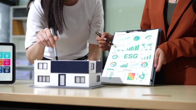 Two women discuss a small house model and sustainability charts during a business meeting about eco friendly building planning