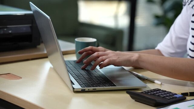 Hands typing on a laptop at a desk in a modern office workspace with a calculator and coffee mug nearby