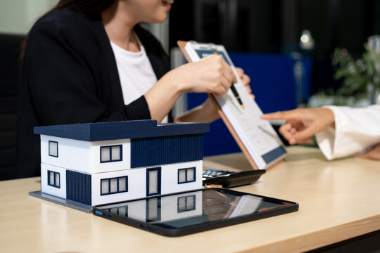 Real estate agent discussing a property offer with a client at a desk, with a house model, clipboard, and tablet in the foreground