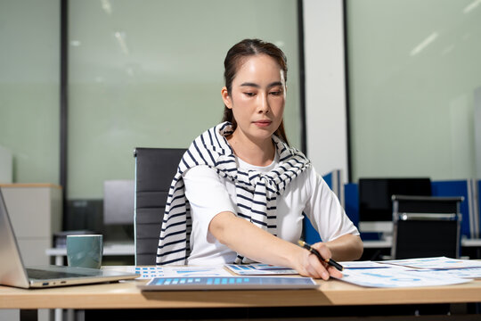 Woman working at office desk, reviewing documents and writing notes beside a laptop in a modern workspace.