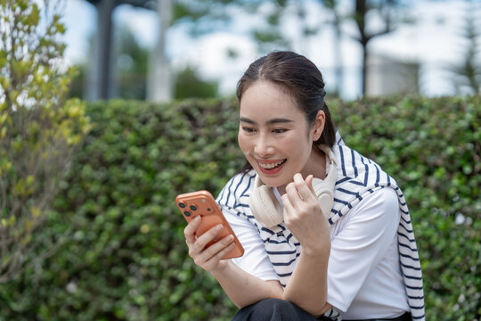 Smiling young woman using a smart phone outdoors with headphones around her neck, looking happy and relaxed in a park