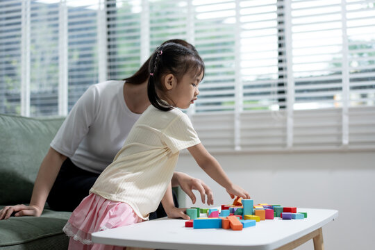 Mother watching her young daughter play with colorful blocks at a white table in a bright living room
