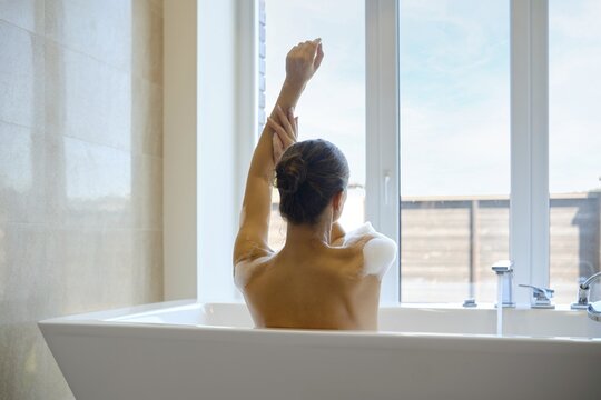 An unrecognizable woman enjoys a tranquil moment in a spacious bathtub while extending her arm. Sunlight streams in through large windows, enhancing the serene atmosphere of the modern bathroom space