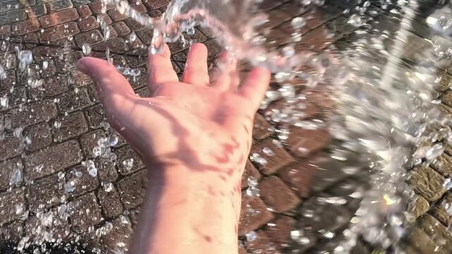 A bare hand rotates under a falling water stream outdoors, turning from palm up to the side as splashes scatter steadily across a wet brick pavement surface in bright daylight.