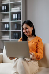 Asian woman feeling happy and smiling while relaxing on a sofa at home. Working or studying using a laptop. Representing remote work. Online learning © David