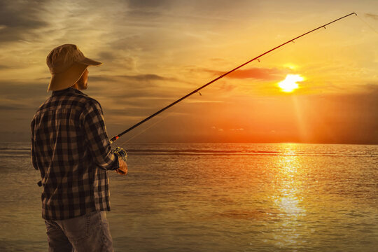 Back view of man wearing hat fishing by ocean at sunset