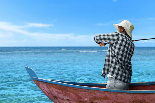 Man wearing hat fishing from wooden boat on ocean under blue sky