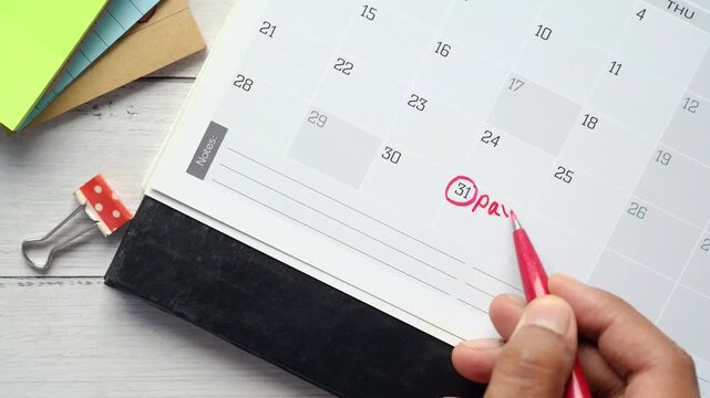 A close up shot of a person marking a calendar with a red pen, circling the 31st and writing pay off debt as a reminder for financial planning and debt repayment