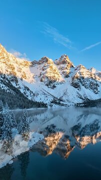 Snowy Mountain Reflection on Winter Lake at Sunrise - A serene winter morning scene unfolds as a vast, calm blue lake perfectly reflects snow-capped mountains illuminated by the golden sunrise.