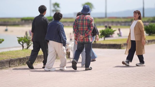 Sunlight warms a paved path beside greenery. People stroll, chat, and pause as they walk. Some lean in, smiling. Others look ahead, relaxed. Lush trees and distant hills frame the scene