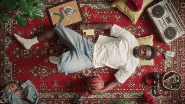 Top view of young Black man lying on red retro carpet, holding basketball and looking at camera, surrounded by 90s analog electronics and classic boombox. Zoom in, video portrait