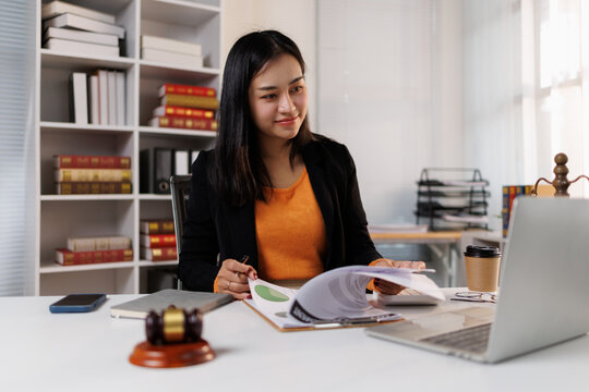 Young businesswoman or lawyer working in a modern office, reviewing legal documents and financial reports with a laptop and gavel on her desk, managing business and justice