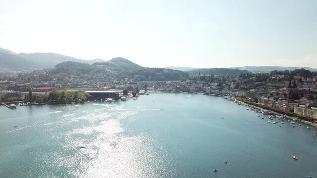 Backlit drone view looks into Lucerne bay toward KKL Luzern, Chapel Bridge and the Reuss outlet with traffic below