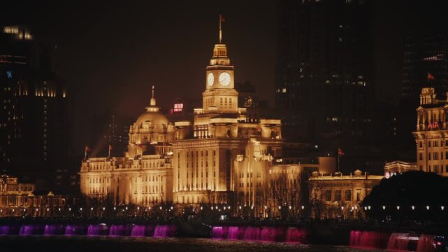 Telephoto night shot of the central Bund heritage facades from across the Huangpu River, Shanghai. The Customs House clock tower dominates centre frame with Chinese flag flying from the top.
