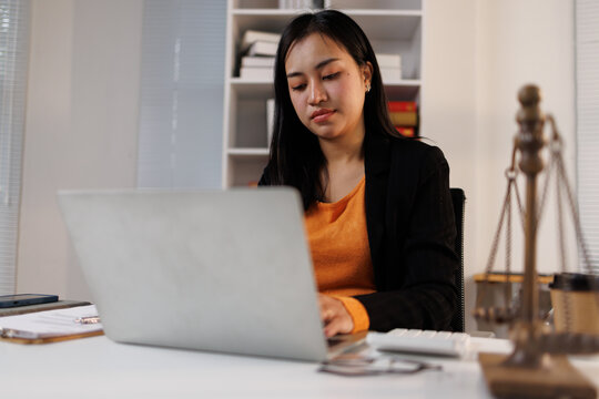 Young asian woman lawyer or attorney using a laptop in a contemporary law office setting, focusing on her work with legal documents and the scales of justice visible on her desk
