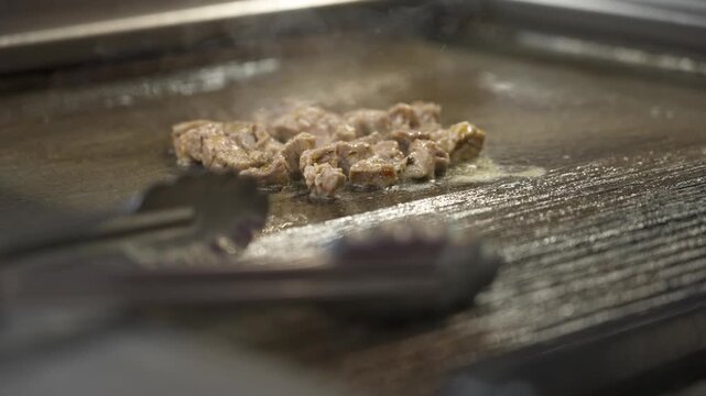 Thin pieces of chicken sizzle in oil on a hot metal griddle as stainless tongs rest in the foreground.