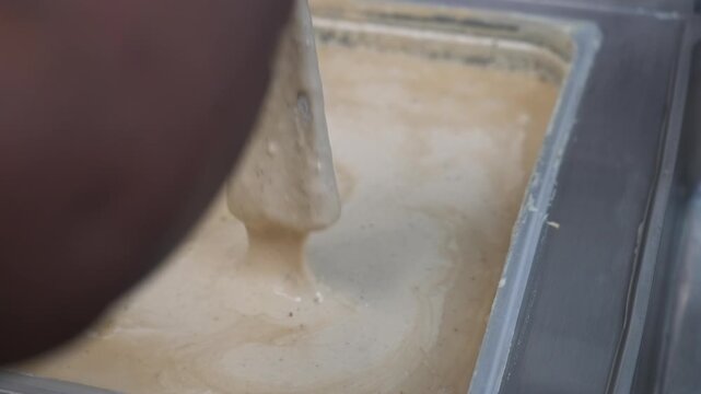 Gloved worker dips a food item into a deep tray of thick seasoned batter in a takeaway store kitchen.