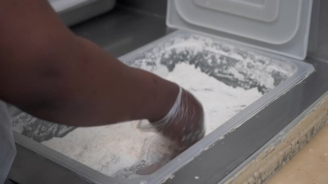 Cook wearing a glove dredges a raw fish fillet in a deep flour tray in a commercial kitchen prep area.
