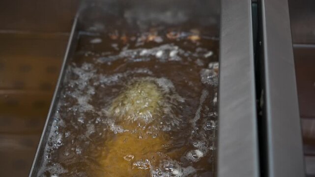 Gloved hand submerges a battered food item into bubbling hot oil inside a stainless steel deep fryer.