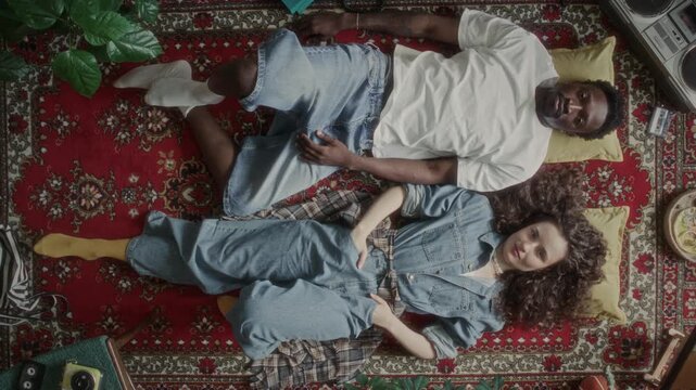 Top view of young Black man and Caucasian woman posing for camera while lying on patterned vintage rug, surrounded by 90s analog electronics and decor. Zoom out shot