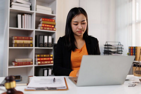 Asian female lawyer working on laptop in modern office, managing legal documents and consulting law books for justice and professional advice